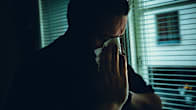 Henkilö seisoo ikkunan edessä pitäen kasvoillaan nenäliinaa hämärässä huoneessa.