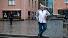 Kalju keski-ikäinen valkoharmaaseen pusakkaan ja farkkuihin pukeutunut mies seisoo sateisessa kaupunkimaisemassa Mikkelissä, nojaa kävelykadun varrella olevaan betonitolppaan.