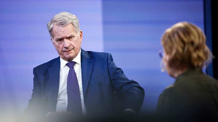 A white-haired man in a blue suit and tie sits in a TV studio speaking to a brunette woman with her back to the camera. 