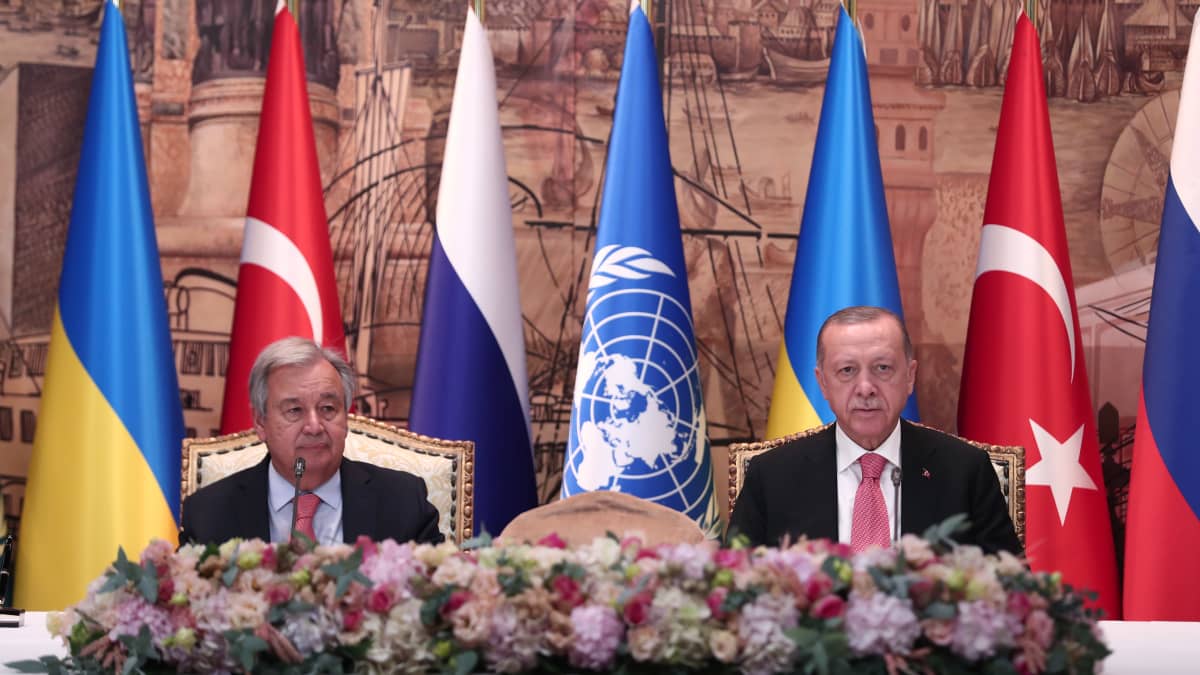 Kaksi pukuun pukeutunutta ihmistä istuu vierekkäin. Etualla kukka-asetelma. Taustalla Ukrainan, Turkin. YK:n ja Venäjän lippuja.