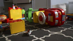 Päällä istuttava lelu auto makaa matolla kellallaan. Lattialla värikkäitä leluja.