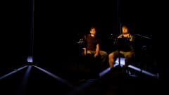 Janne Suutarinen ja Markus Serala istuvat tuoleilla pimeällä lavalla. Edessään heillä on mikit ja nuottitelineet. Vain nuottitelineiden valot valaisevat heitä.