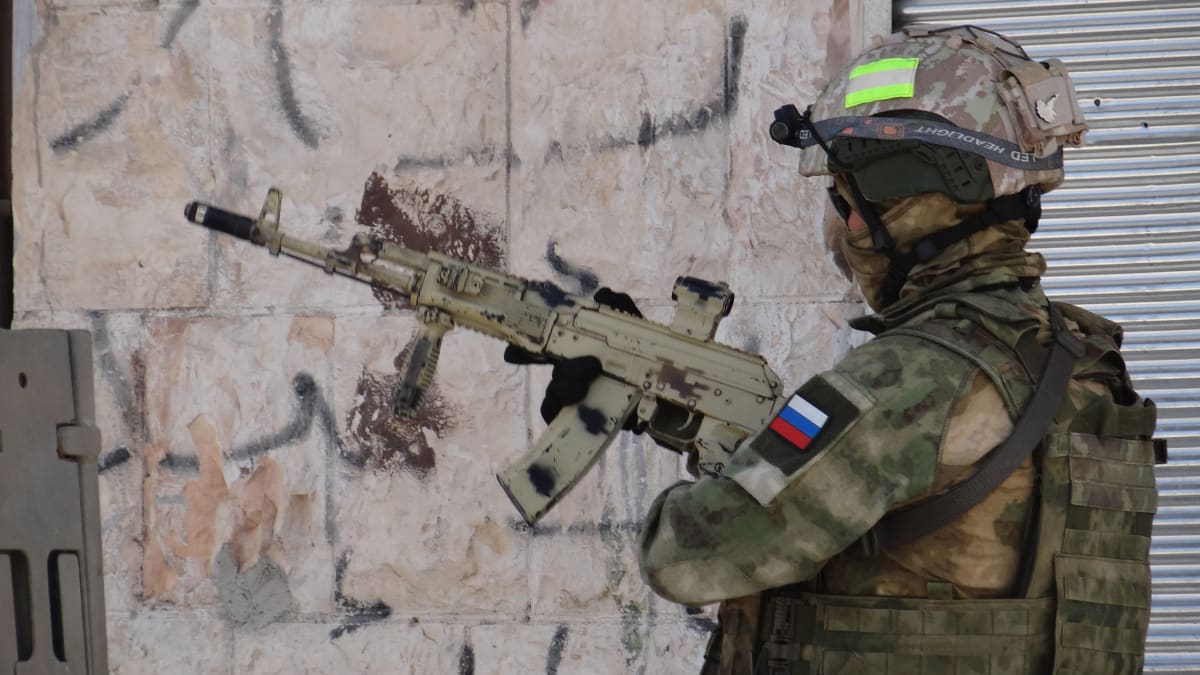 Venäläinen sotilas rynnäkkökiväärin kanssa, hihassa Venäjän lippu, taustalla seinä jossa ilmeisesti arabian kielisiä graffiteja. 