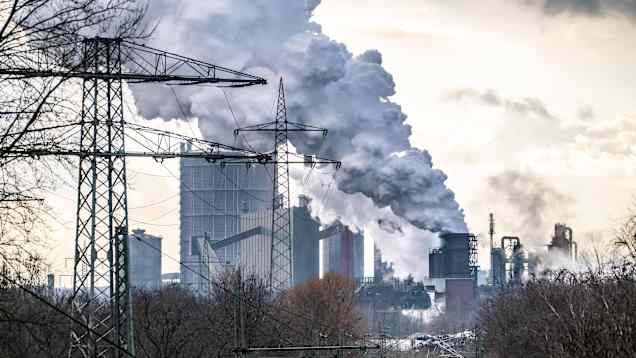 Rök väller fram mot himlen från höga fabriksskorstenar, i förgrunden kraftledningar och en tågbana.