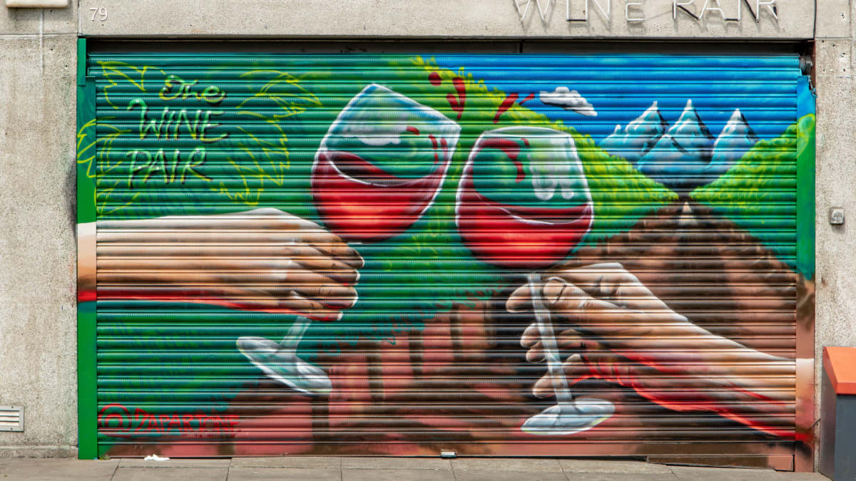 Katutaidetta Dublinissa: The Wine Pair -viinikaupan ja -baarin ikkuna on peitetty metalliverholla, johon on maalattu kaksi punaviinilasilla kilistelevää kättä.