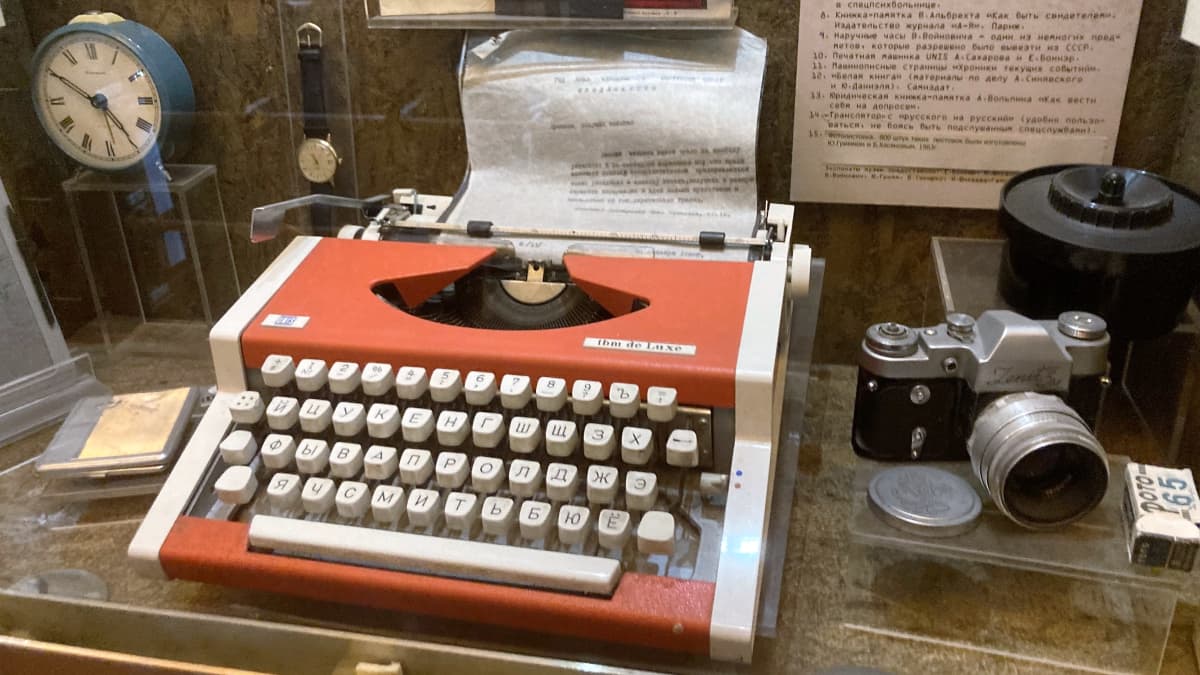 Näyttelyvitriinissä on kirjoituskone, jossa on kyrilliset kirjaimet. Koneen vieressä on kamera. Hyllyköllä on myös muun muassa vanha herätyskello ja rannekello.