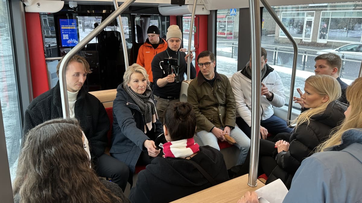 Ratikkavaunussa istuu penkeillä ehdokkaita, keskellä istuu mikrofoni kädessä Katri Tapola. Taustalla näkyy lähetystä kännykällä kuvaava Oskari Räisänen. 