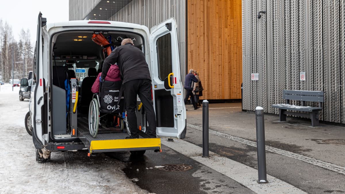Taksinkuljettaja auttaa pyörätuolilla kulkevaa asiakasta taksin kyytiin Sairaala Novan pihassa.