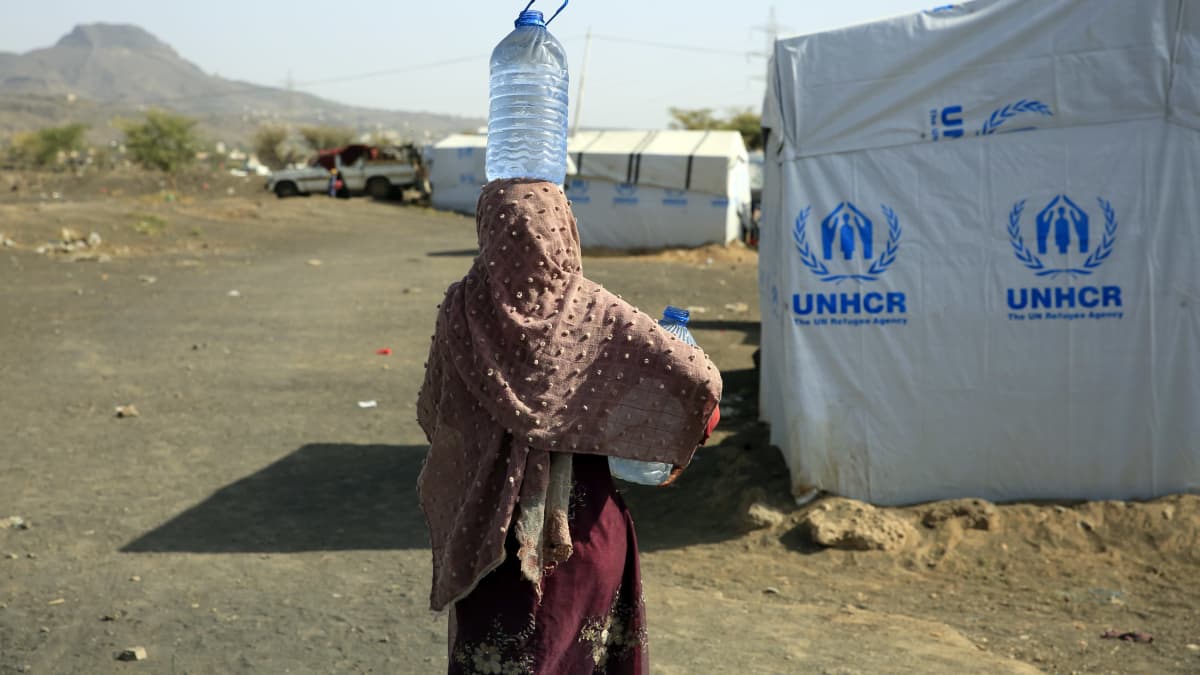 Nainen kantaa vesipulloa pään päällä UNHCR:n teltan edessä. 