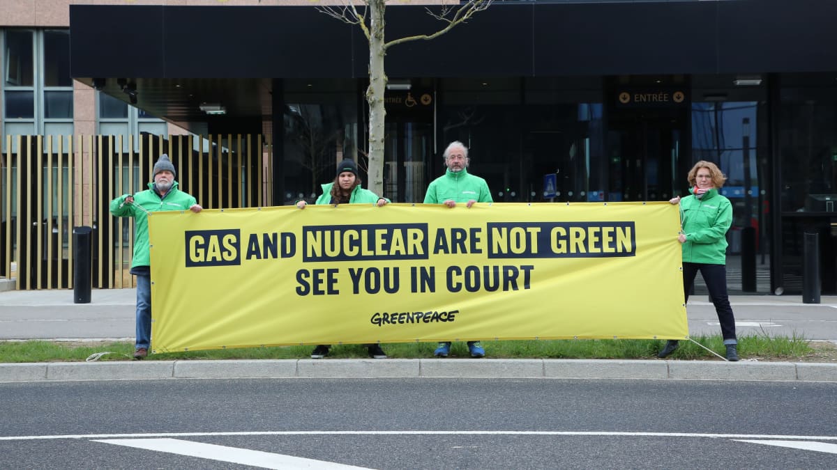 Neljä vihreään Greenpeace-takkiin puekutunutta ihmistä pitää käsissään keltaista lakanaa, jossa lukee "Gas and Nuclear are not green. See you in court".