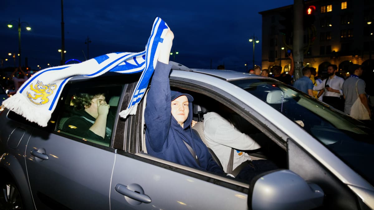 Huuhkaja fani istuu autossa ja suomen lippu liehuu.