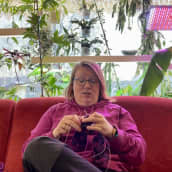 Mari Liedes neuloo käsityötä punaisella sohvalla, jonka takana on paljon viherkasveja.