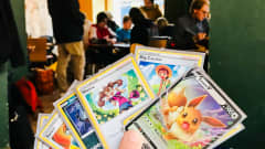 Seitsemän erilaista Pokemon korttia henkilön kädessä. Taustalla näkyy muita Pokemon pelaajia.