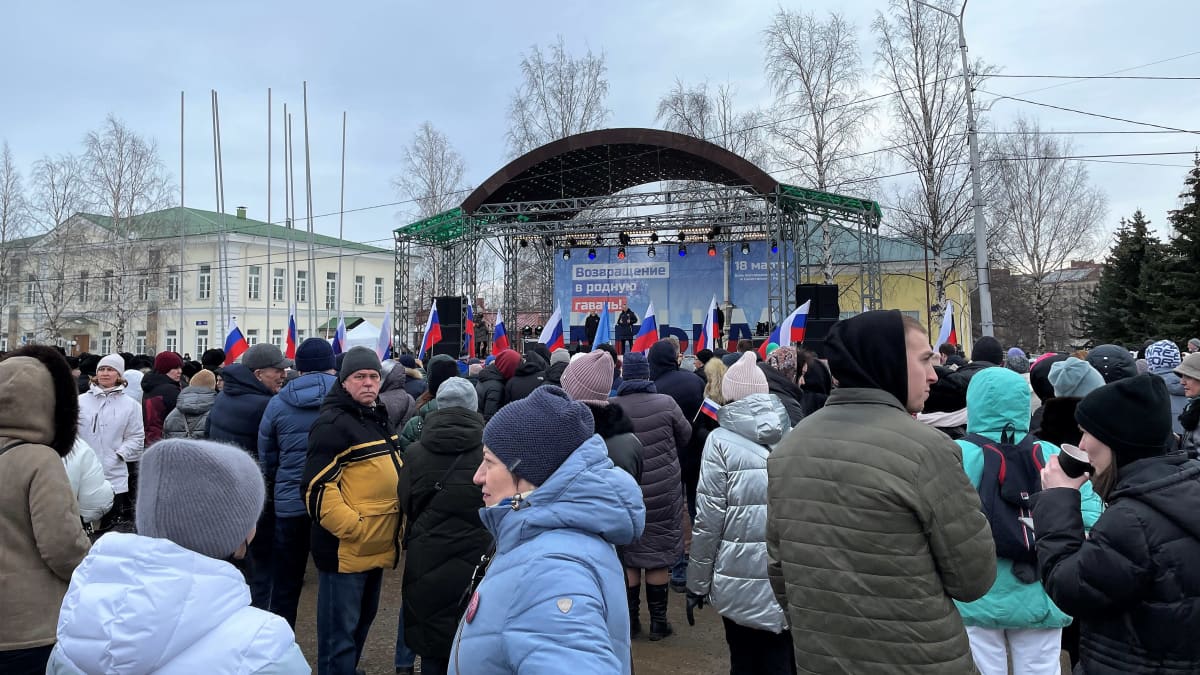 Talvivaatteisiin pukeutunut väkijoukko seisoo juhlalavan edustalla. Lavan Venäjän lipun värein koristellussa taustajulisteessa lukee venäjäksi: Paluu kotisatamaan.