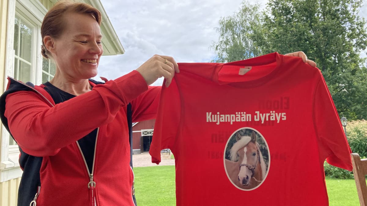 Nainen pitelee käsissään punaista paitaa, jossa on hevosen kuva.