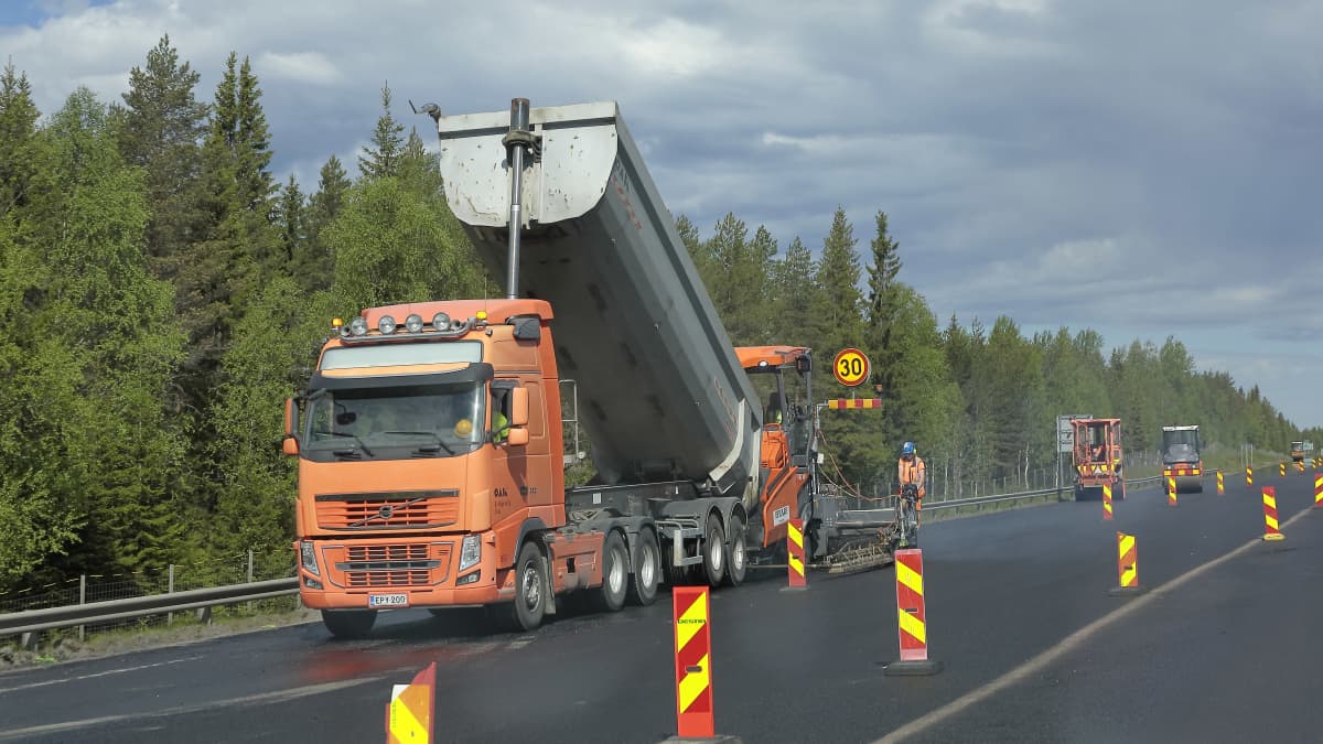 Oranssi kuorma-auto lava pystyssä upouudella asfaltilla, varoitusmerkkejä ja huomioasuisia asfalttityöntekijöitä seisoo tiellä.