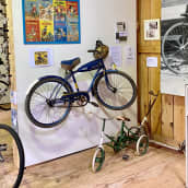 Polkupyöriä, Aku Ankan sinininen nimikkopyörä 1950-luvulta kuvan keskellä seinälle nostettuna. Pyörän etulokasuojassa on Aku Ankan pää ja sen sarvessa roikkuu keltainen, Aku Ankka-kuvioinen kypärä. 
