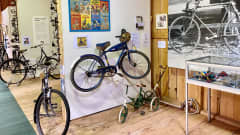 Polkupyöriä, Aku Ankan sinininen nimikkopyörä 1950-luvulta kuvan keskellä seinälle nostettuna. Pyörän etulokasuojassa on Aku Ankan pää ja sen sarvessa roikkuu keltainen, Aku Ankka-kuvioinen kypärä. 
