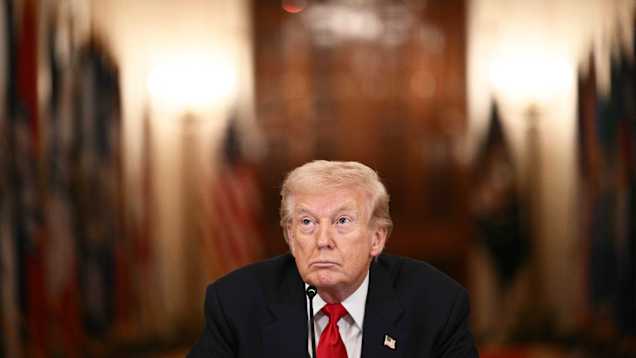 Donald Trump sitter vid ett bord med en mikrofon framför sig. I bakgrunden skymtar suddiga flaggor.