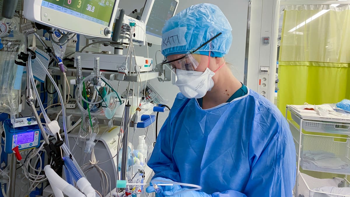 Sairaanhoitaja sinisessä suoja-asussa monitorien ja laitteistojen edessä tekemässä hoitotoimenpiteitä teho-osastolla-