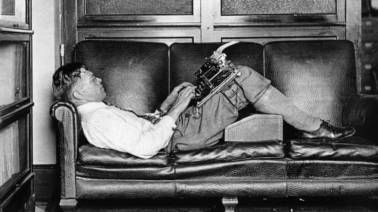 Mies makaa nahkasohvalla ja kirjoittaa kirjoituskoneella joka on hänen jalkojensa päällä.