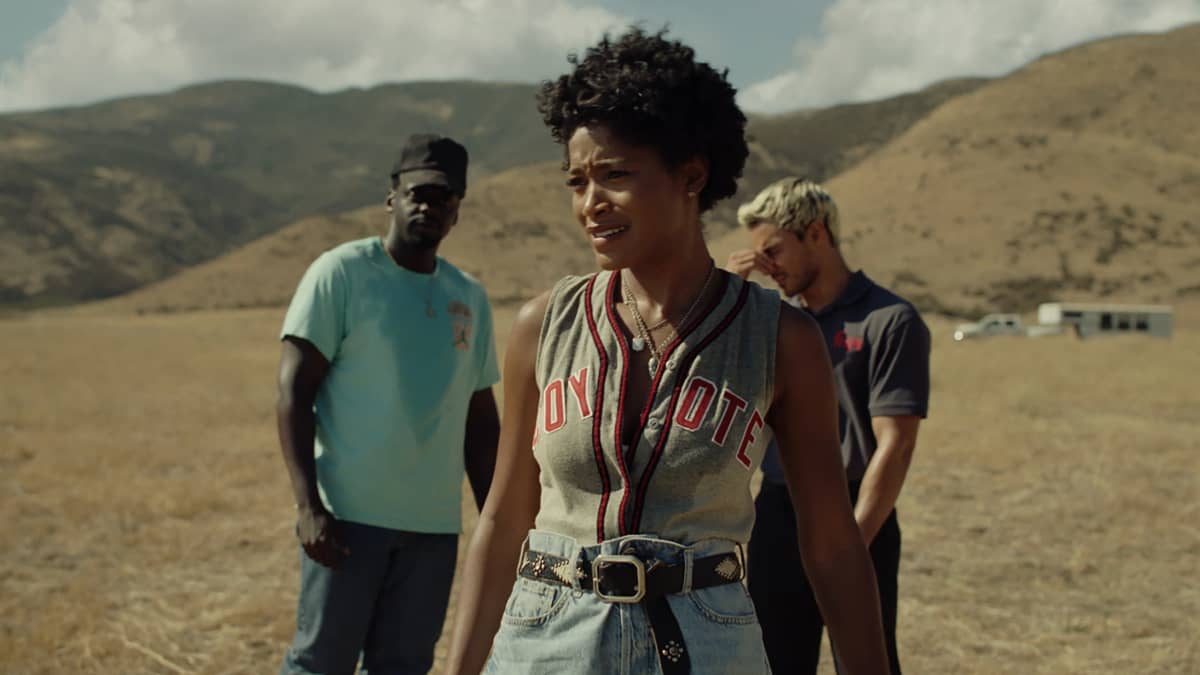 Musta nainen aavikolla baseball-t-paidassa, taustalla kaksi miestä.