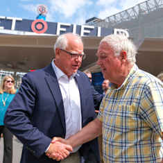 Tim Walz kättelee ruutupaitaista, harmaatukkaista miestä, taustalla stadionrakennelmia.