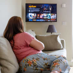 A woman sits on a sofa looking for something to watch Netflix on a television on the wall.
