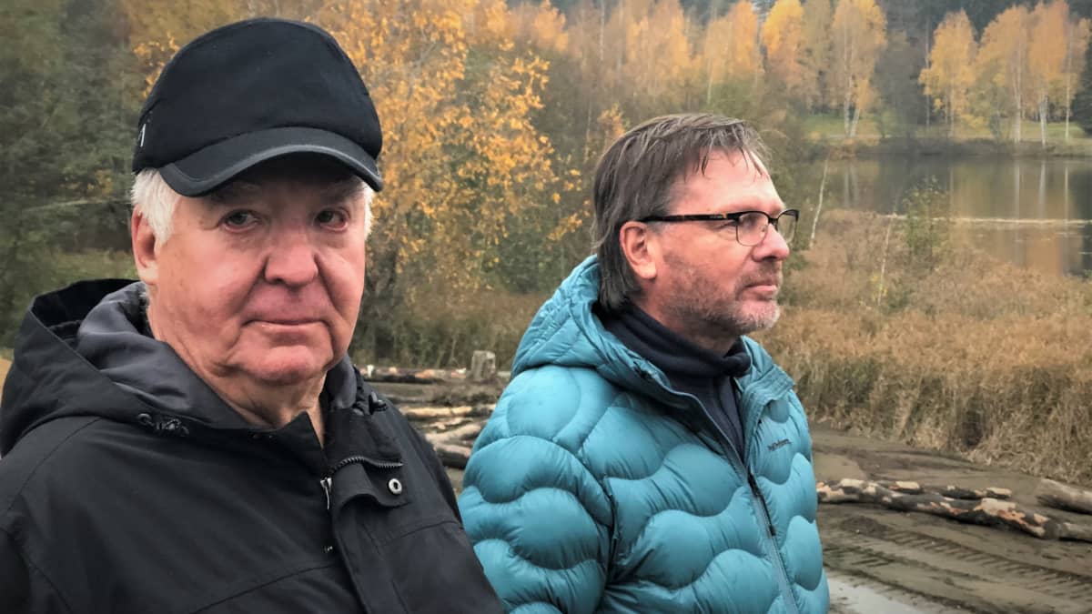 Kaksi miestä seisoo vierekkäin, toinen katsoo kameraan, taustalla järvi ja syksyisiä puita. 