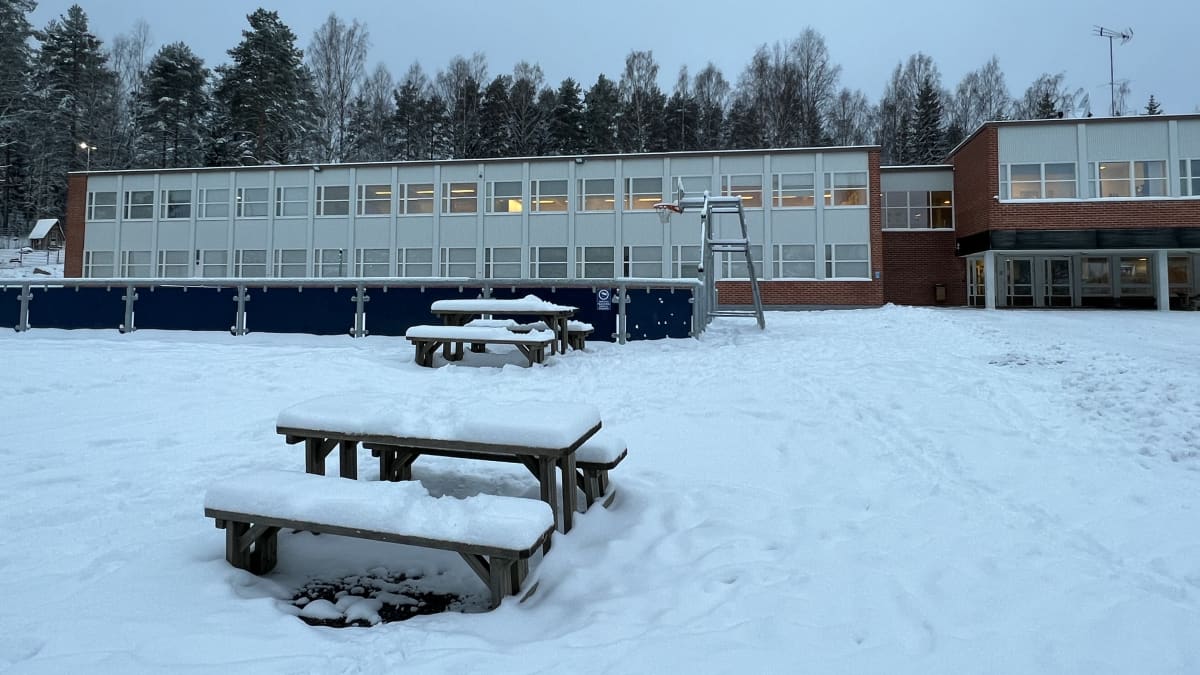 Lumen peittämä koulupiha, jossa on muutamia puisia penkkejä ja pöytiä. Taustalla kaksikerroksinen tiilitalo.