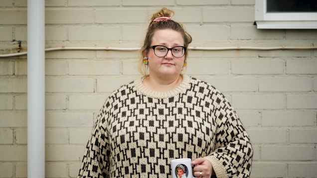 En person i mönstrad tröja håller en mugg framför en ljus tegelvägg.