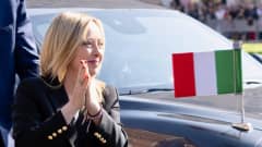 Mustajakkuinen vaalea nainen painaa kämmenensä yhteen mustan auton edessä ja hymyilee oikealle päin. Taustalla auton konepellillä on Italian lippu.