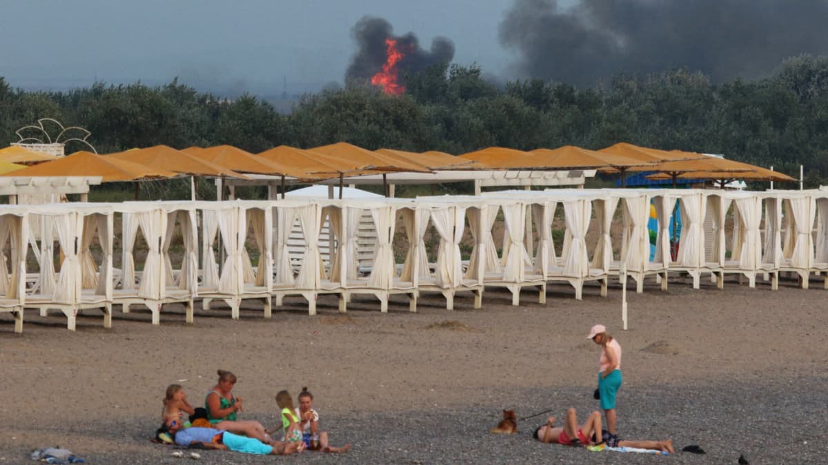 Venäläiset turistit viettävät aikaa rannalla Krimin niemimaalla. Taustalla palaa ja savua nousee ilmaan.
