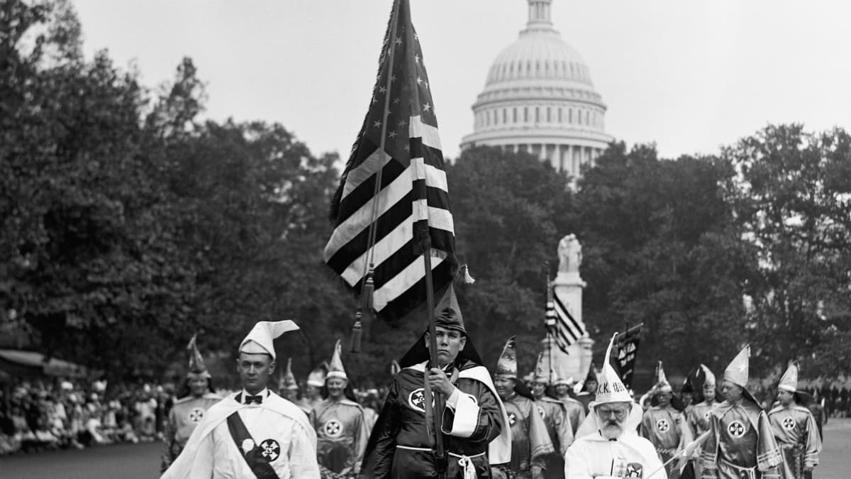Ku Klux Klanin kaapuihin pukeutuneet miehet marssivat pitkin katua. Taustalla näkyy kongressin rakennus Capitol Hillillä.
