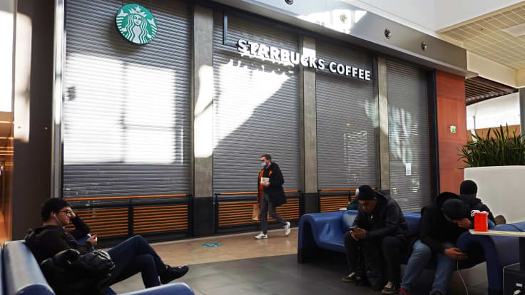 Suljettu Starbucksin kahvila. Etualalla istuu ihmisiä puhelimiinsa syventyneinä, yksi henkilö kävelee kahvilan peitettyjen ikkunoiden ohi.
