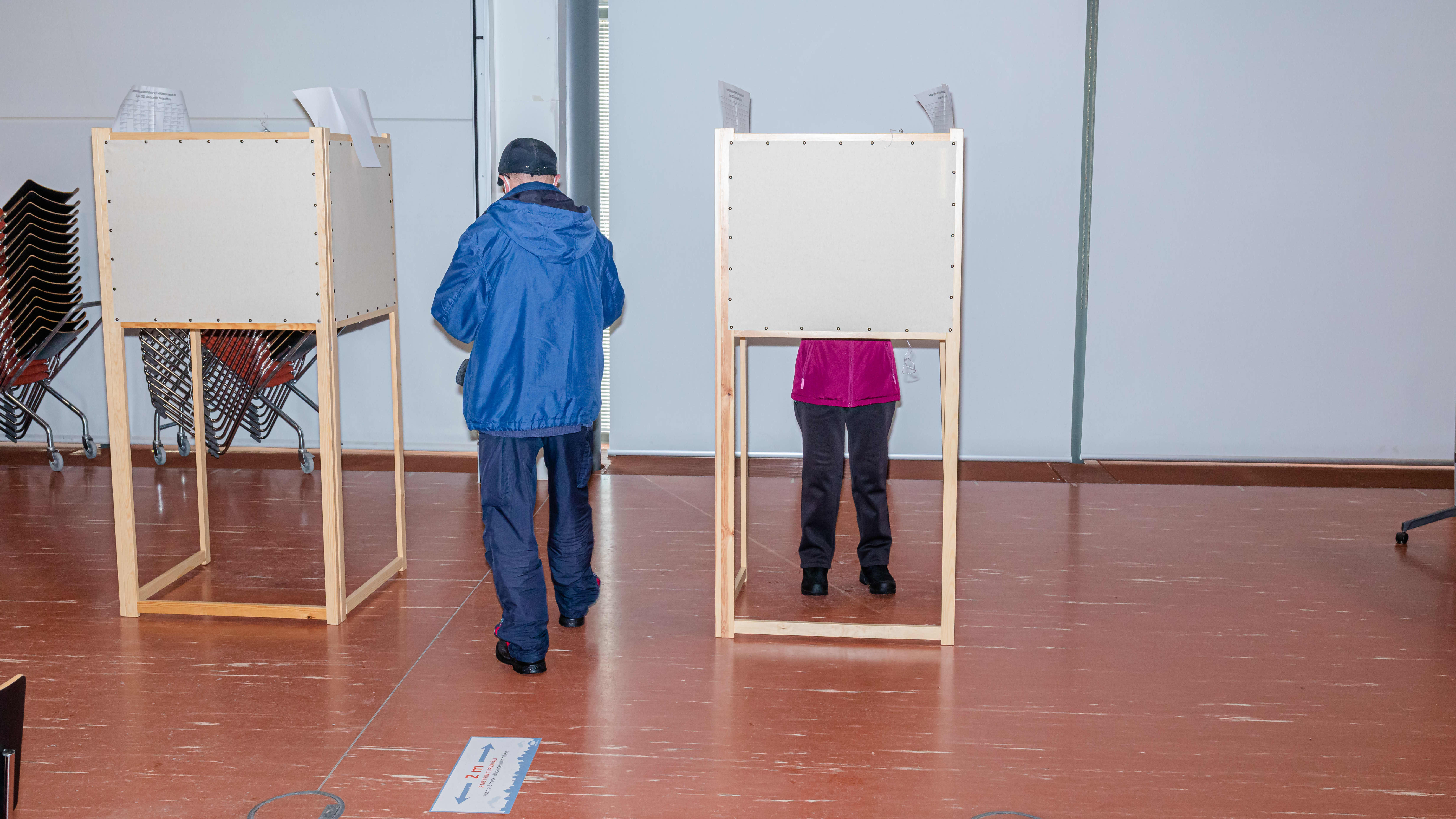 Kaksi äänestyskoppia huoneessa, toisessa kopissa on äänestäjä, joka on pukeutunut punaiseen takkiin, toiseen äänestyskoppiin on matkalla henkilö jolla on sininen takki.