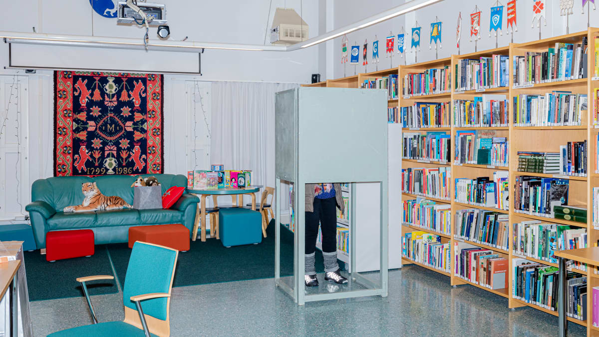 Aluevaalien ennakkoäänestyspaikka pienessä kirjastohuoneessa, kirjahyllyt täyttävät yhden seinistä, keskellä harmaansininen äänestyskoppi, taka-alalla sohva jossa pehmoeläimiä, kuten tiikeri.