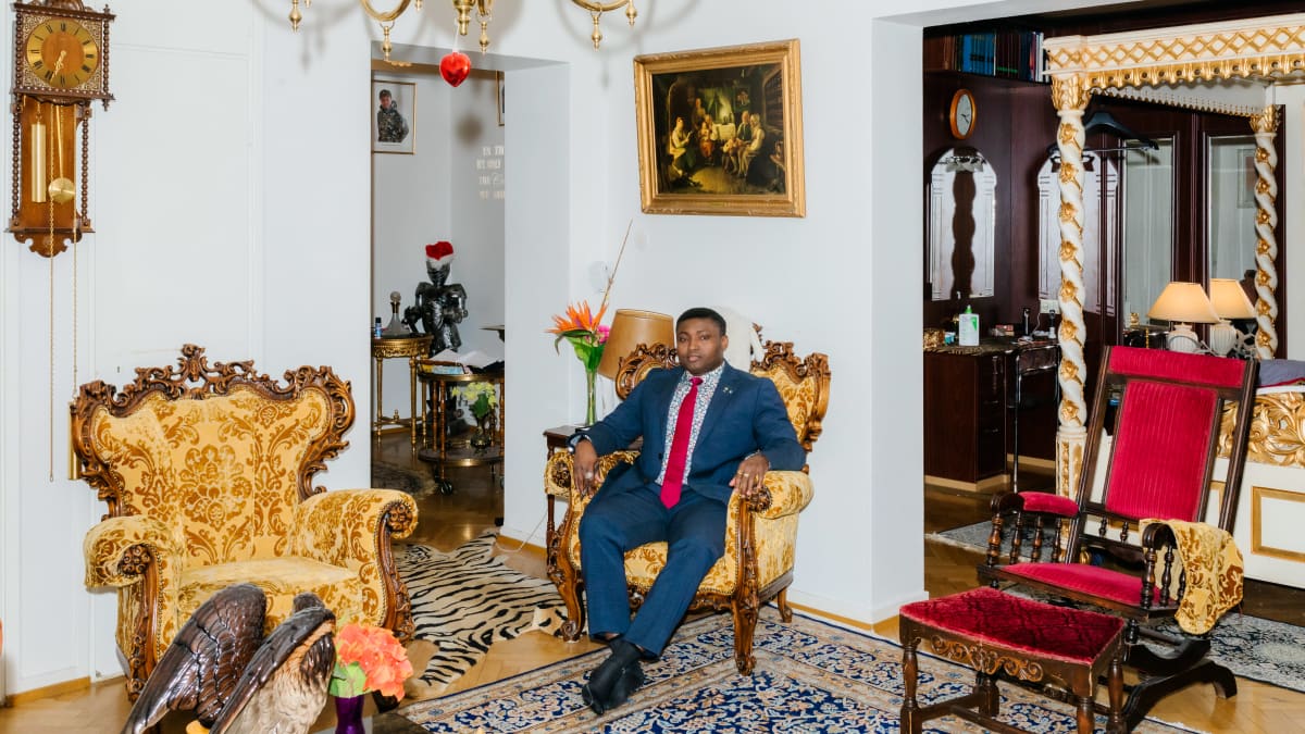 Simon Ekpa is sitting in an armchair in the living room, the inscription "Dare to dream big" can be seen on the wall.