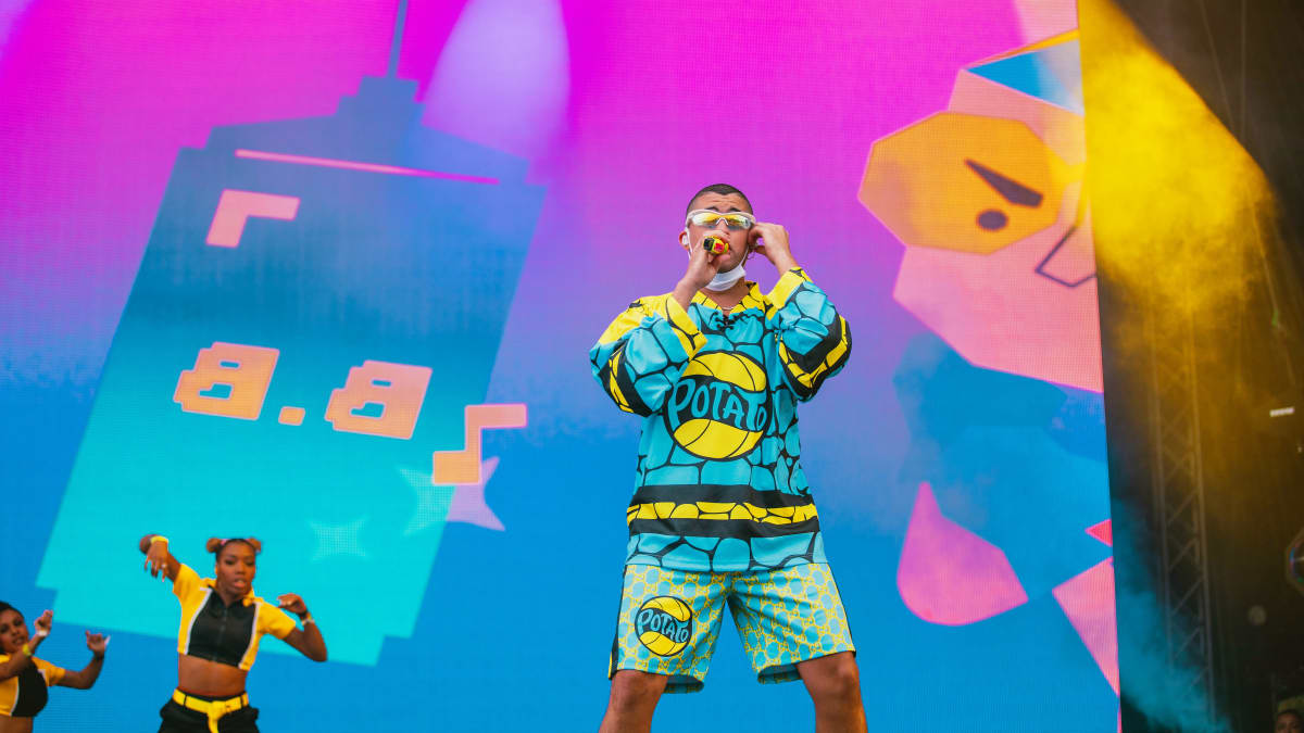 Siniseen kuviolliseen asuun puketunut portugalilaisartisti Bad Bunny laulaa mikkiin Ruisrockin Rantalavalla. Taustlla näkyy kaksi keltamustaan pukeutunutta taustatanssijaa ja värikkäät videovisuaalit.