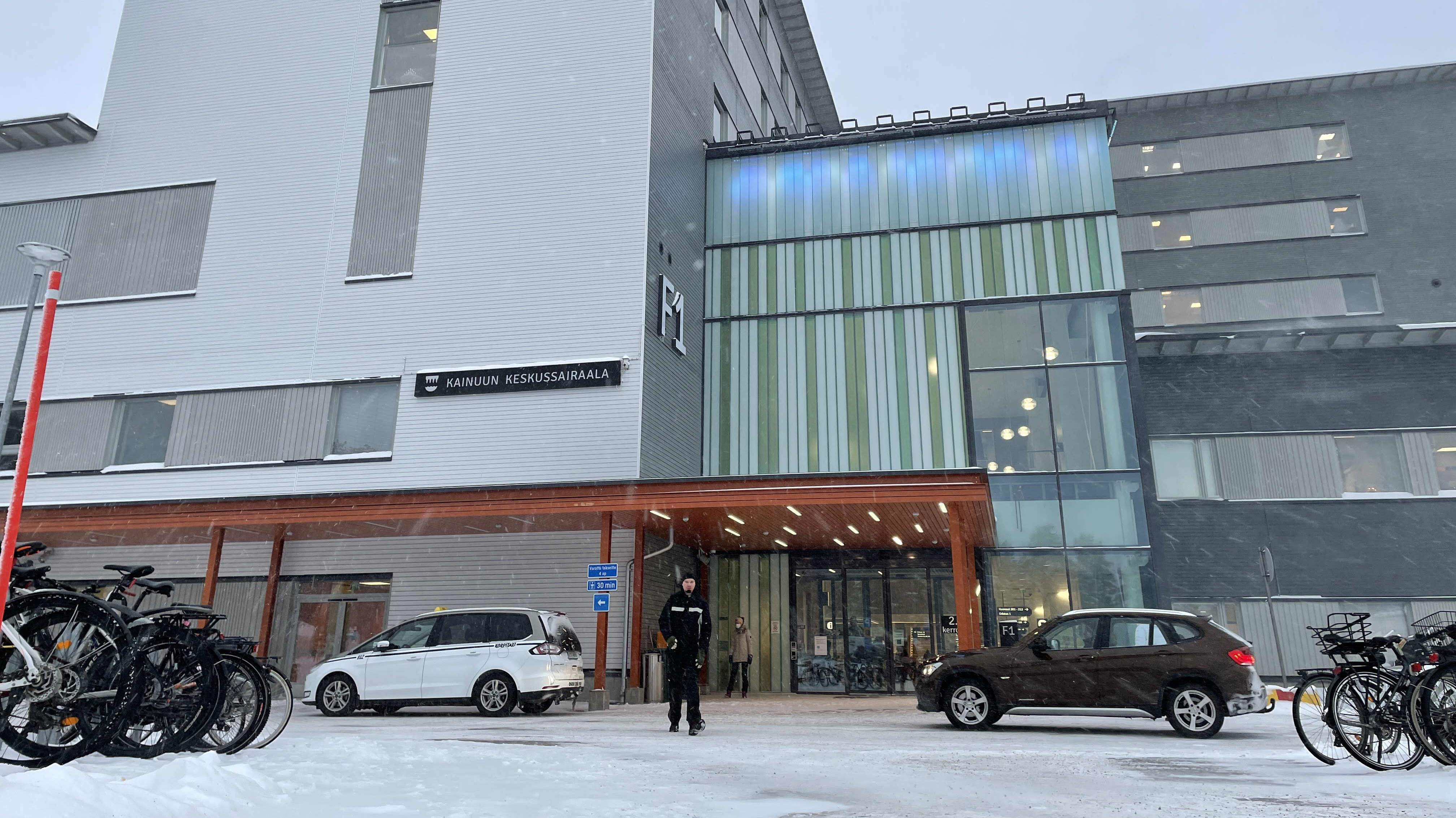 Kainuun keskussairaalan pääsisäänkäynti lumisella kelillä. Mustiin pukeutunut mies kävelee tien yli poispäin sairaalasta.