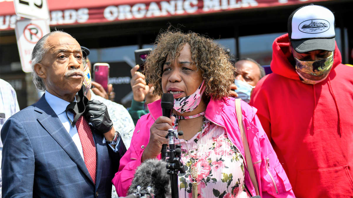 Eric Garnerin äiti Gwen Carr puhuu mikrofoniin. Al Sharpton kuuntelee vakavana vieressä. Taustalla näkyy väkijoukkoa ja elintarvikekaupan julkisivua.