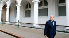 Vladimir Putin kävelee tummansinisessä puvussa ja kravatissa pylväskäytävämn edustalla ja katselee ylös kuvan vasempaan reunaan.