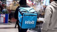 A person with a black hoodie and a large cubical blue backpack saying Wolt Partner seen from behind in a shopping mall.