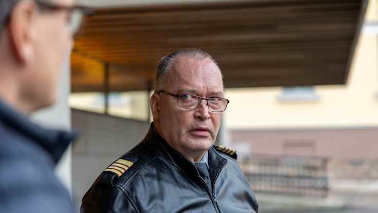 A older man wearing glasses and a leather military jacket seen in half-profile, speaking with a serious expression  to another man in front of a building.