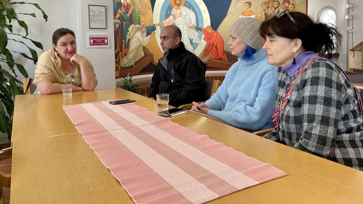 Ihmisiä pöydän ympärillä huoneessa, jonka seinällä on ortodoksien fresko. 