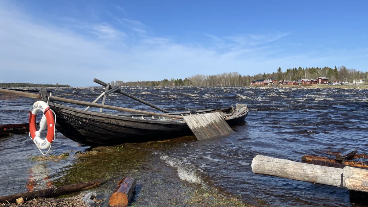 Vanha puuvene jokirannassa ja taustalla koski kuohuu.