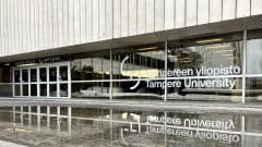 Tampereen  yliopiston sisäänkäynti. Lasi-ikkunoissa teksti "Tampereen  yliopisto - Tampere University". Rakennuksen edessä vesilammikko. 