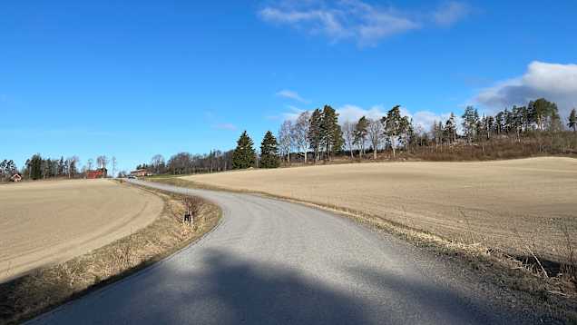 En landsväg som slingrar sig genom ett öppet jordbrukslandskap under en klar blå himmel.