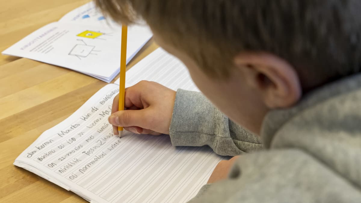 Poika koulussa äidinkielentunnilla kirjoittaa lyijykynällä tarinaa vihkoon tekstaten.