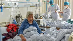 Kaksi koronapotilasta kiinalaisessa sairaalassa. Edessä istuu vuoteessaan vanha nainen, jolla on happiviikset. Takana on kaksi suojavarusteisiin pukeutunutta hoitajaa toisen potilaan vuoteen ääressä. 